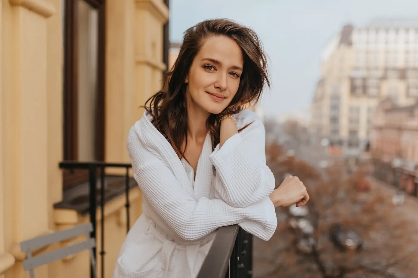 A rested, makeup-free woman in a bathrobe looks at the camera while posing on an open balcony.