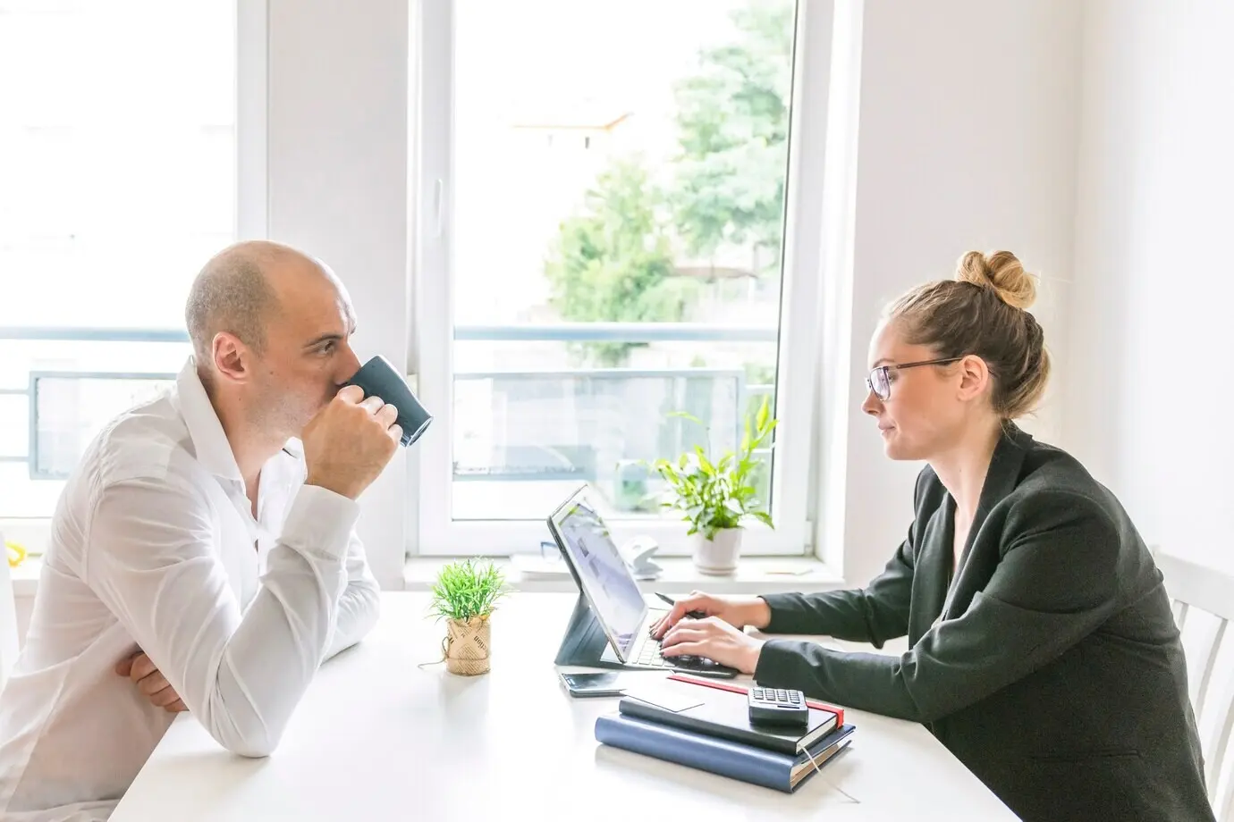 A businessman drinks coffee while watching his partner work on a laptop.
